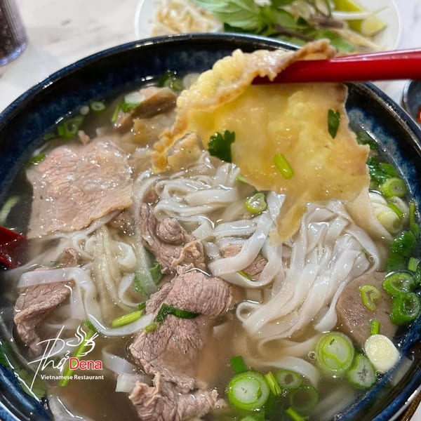 Close-up of Pho Dena's Vietnamese pho with beef, noodles, and green onions, with a piece of tripe held by red chopsticks.