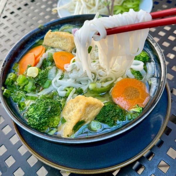 Chopsticks lifting flat rice noodles from a bowl of Pho Dena's vegetarian pho with tofu and vegetables.